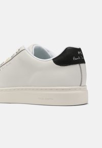 Białe skórzane sneakersy z czarną kontrą na pięcie, wytłoczonym logo i kontrastującym przeszyciem. Charakteryzują się minimalistycznym designem z subtelnymi akcentami kolorystycznymi.