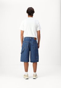 Shorts en denim bleu, dotés de multiples poches, d'une étiquette en cuir à l'arrière et d'un ourlet brut. Associés à un t-shirt blanc uni et des baskets.