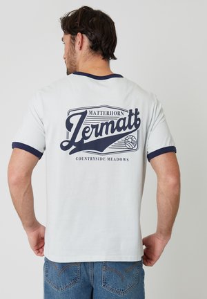 Mand iført hvid T-shirt med marineblå kant, ryggen viser teksten "Matterhorn Zermatt Countryside Meadows" og et bjergmotiv.