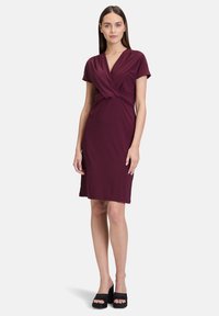 Bordeaux knielange jurk met een wikkeldesign, korte mouwen, zachte stretchstof en een geknoopte details in de taille. Geraadpleegd met zwarte hoge hakken sandalen.
