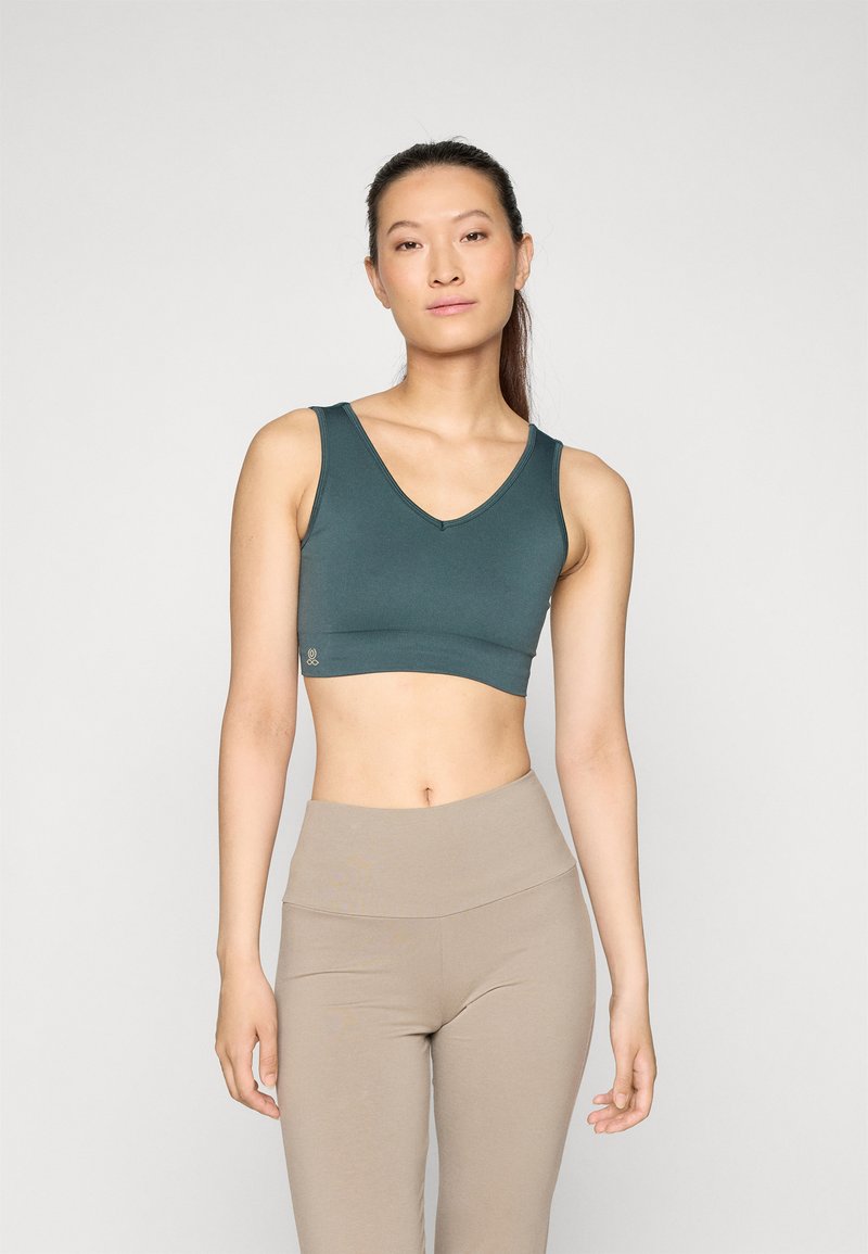 Sujetador deportivo de pico en color teal, hecho de tela elástica, con una forma ajustada y tirantes anchos. Combinado con leggings beige de tiro alto.
