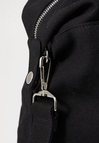 Bolsa de lona negra con cremalleras plateadas, botón de presión y un clip metálico. Cuenta con una correa resistente y tela texturizada.