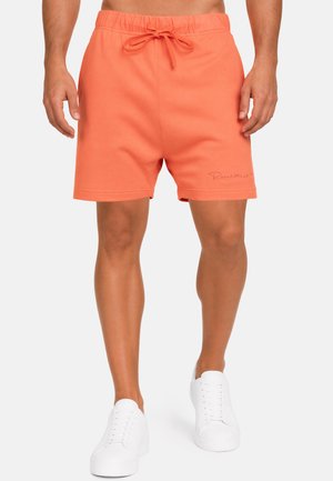 Mann trägt korallenorange Shorts mit Kordelzug und einem dezenten Logo am linken Bein sowie weiße Freizeitsneaker und steht vor weißem Hintergrund.