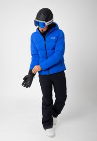 Blå isolerad jacka med huva, svarta byxor och vita sneakers. Bär en hjälm och goggles, justerar en svart handske.