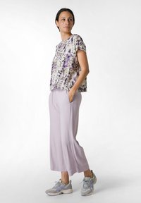 Ljuslila vida byxor i kombination med en kortärmad, mönstrad topp i lila och kremfärger. Modellen står med händerna i fickorna.