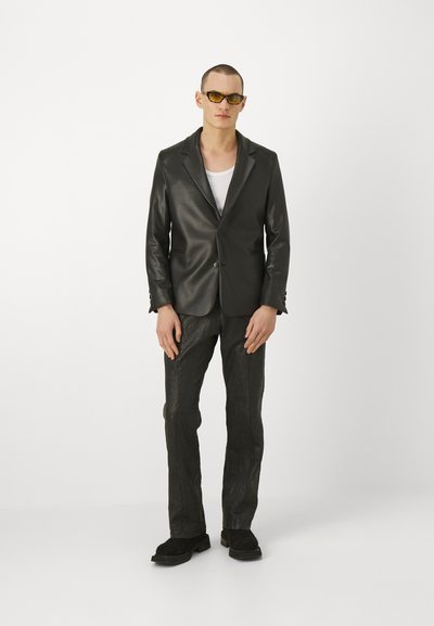 Čierny kožený blazer s jedným gombíkom, v kombinácii s textúrovanými čiernymi nohavicami. Model nosí slnečné okuliare a biely tielko pod ním.