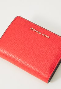 Portefeuille en cuir rouge avec une surface texturée ; présente un logo doré "MICHAEL KORS" et un bord noir sur les côtés. Forme rectangulaire.