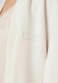Hellbeige Cardigan mit weicher Textur. Mit einem gestickten Logo 'CCDK COPENHAGEN' auf dem Ärmel. Einfaches, offenes Design.