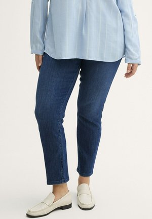 Chemise à manches longues bleu clair, assortie à un jean en denim bleu foncé et des mocassins blancs. La tenue présente une coupe décontractée et un design simple.