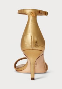 Sandálias de salto douradas com textura de crocodilo, faixa no tornozelo e bico fino. O salto é esguio e tem cerca de sete centímetros de altura.