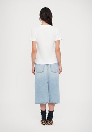 Femme aux cheveux foncés portant un haut blanc à manches courtes, une jupe midi en jean bleu clair, des chaussettes noires et des chaussures à motif léopard, debout de dos.
