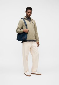 Mann trägt beigen Sweatshirt mit halbem Reißverschluss, weiße Hose, weiße Sneaker, trägt marineblaue Umhängetasche, steht vor einfarbigem Hintergrund.