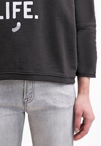 Felpa nera con testo bianco "LIFE." e una piccola grafica. Il modello indossa jeans grigi chiari. Tessuto liscio con polsini a costine.
