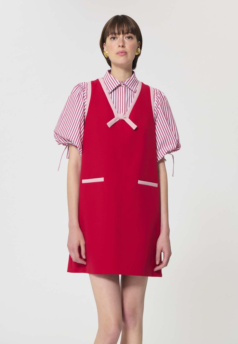 Femme portant une robe rouge sans manches avec un bord rose, par-dessus un chemisier à manches bouffantes rayé rouge et blanc, debout devant un fond uni.
