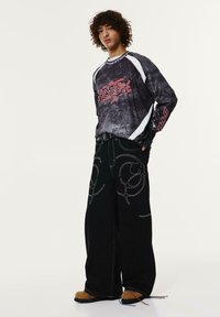 Črne široke hlače z belimi abstraktnimi šivi, v kombinaciji z dolgim zgornjim delom temne barve, ki ima rdeče grafike.