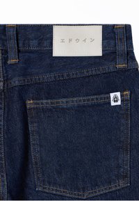 Poche arrière de jean en denim bleu foncé avec des surpiqûres orange contrastantes et une étiquette rectangulaire blanche avec un texte japonais au-dessus de la poche.