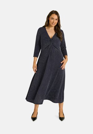Femme debout portant une longue robe sombre scintillante avec des manches trois-quarts et des chaussures noires à bout pointu sur un fond blanc uni.