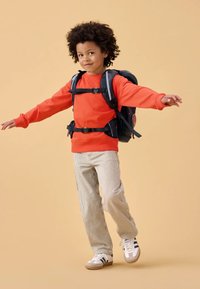 Felpa arancione, pantaloni a righe beige, sneakers bianche con strisce marine e uno zaino nero con spallacci regolabili e dettagli laterali.