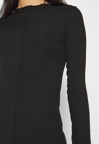 Femme portant un top noir côtelé ajusté à manches longues avec une couture centrale et un col rond légèrement froncé.