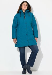 Tealfärgad vattentät parka med huva, knäppning med knapp och dragkedja, sidofickor och en avslappnad passform, kombinerad med blå jeans och svarta stövlar.