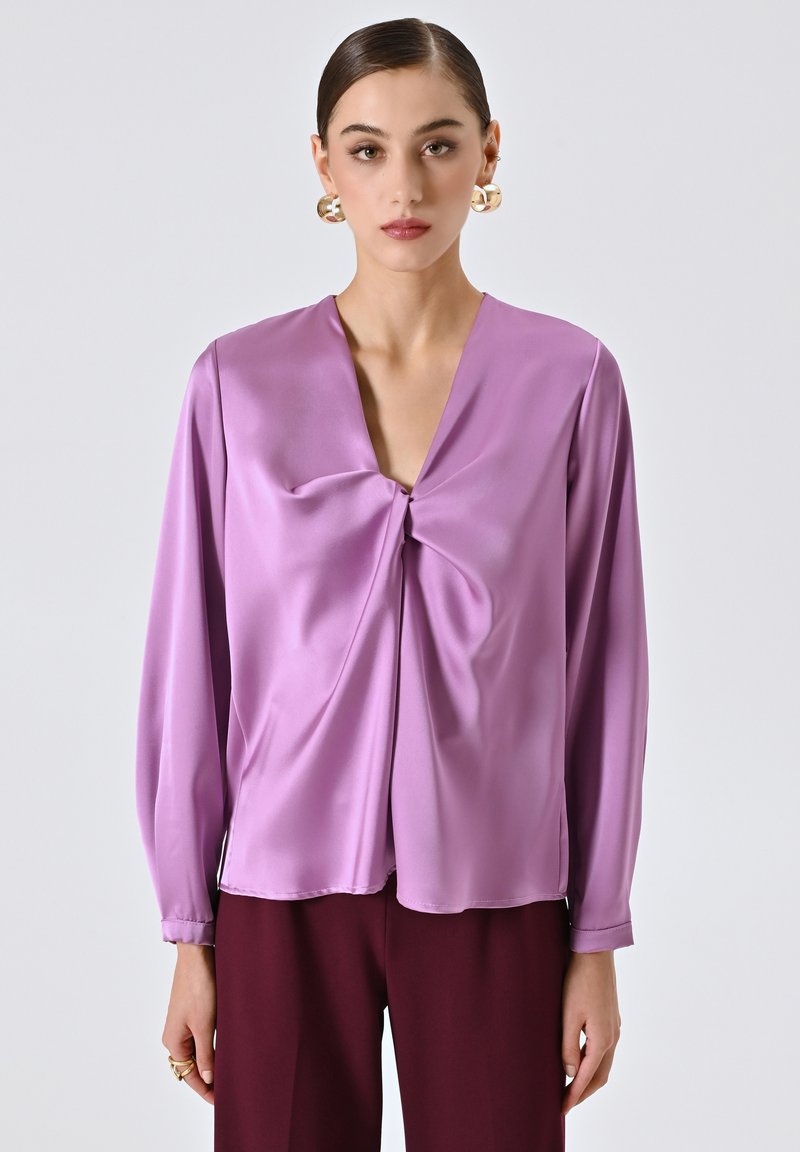 Donna che indossa una blusa lunga a maniche lunghe di un brillante colore lavanda con un dettaglio centrale attorcigliato e pantaloni di un forte bordeaux scuro, accessoriata con orecchini a cerchio dorati.