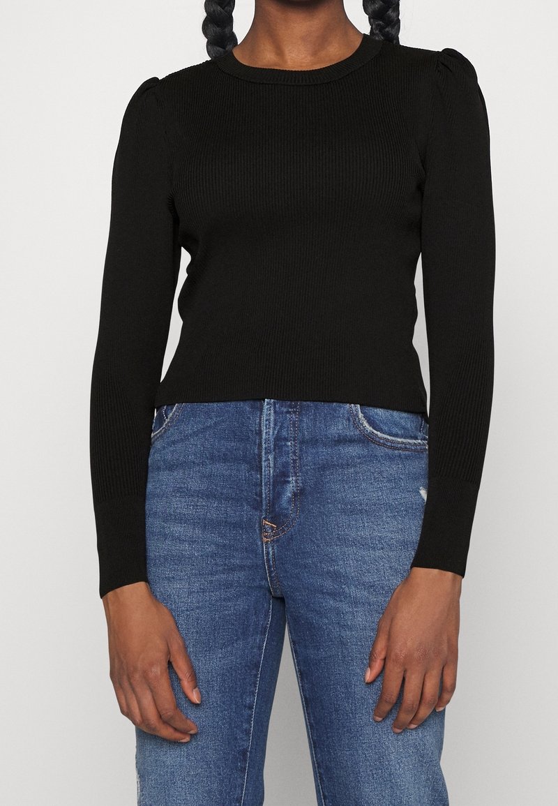 Pull noir côtelé à manches longues avec un col rond et des épaules légèrement bouffantes, associé à un jean en denim bleu.