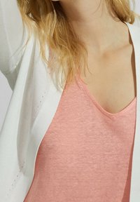 Valkoinen avoin neulottu neuletakki vaaleanpunaisen v-pääntien topin päällä. Kankaat näyttävät pehmeiltä, hieman teksturoituneilta ja rennolta istuvalta.