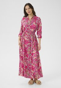 Virágmintás, élénk rózsaszín maxi ruha, hosszú ujakkal, megkötős derékkal és lágy esésű kialakítással. Fonott szandálokkal párosítva.