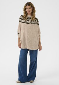 Brede gebreide trui in beige met een zwart geometrisch patroon aan de schouders, gecombineerd met brede blauwe jeans en lichtbeige schoenen. Soepele textuur.