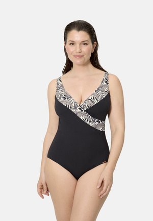 Mujer con el cabello mojado peinado hacia atrás, que lleva un bañador negro de una sola pieza con ribetes florales en beige y negro a contraste, posando frente a un fondo liso.