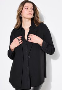 Svart oversized skjorta med knappar, mjuk textur, långa ärmar och skräddarsydd krage. Modellen bär flera silverringar.