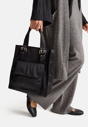 Bolso tote negro hecho de material suave, con doble asa, herrajes plateados y un bolsillo frontal. Combinado con un atuendo de punto gris y zapatos planos negros.