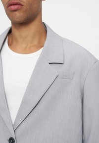 Jeune personne portant un blazer gris clair par-dessus un t-shirt blanc à col rond, coupé au niveau du cou et du haut de la poitrine.