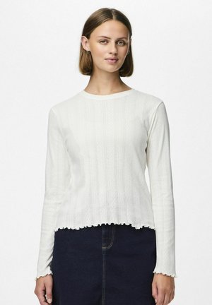 White long-sleeve shirt with ribbed texture, scalloped hem, and rounded neckline. Worn with a blue denim skirt, features a fitted design.