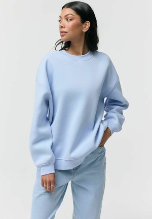 Kobieta o ciemnych włosach, ubrana w jasnoniebieską, oversize'ową bluzę i jasnoniebieskie dżinsy, stojąca z jedną ręką w kieszeni na białym tle.