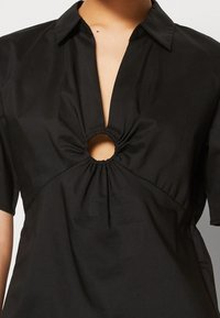 Mustapaita, jossa on kauluskenttä, lyhyet hihat ja keskellä pyöreä aukko. Kangas on sileä ja hieman kiiltävä, ja se kerääntyy vyötärön ympärille.