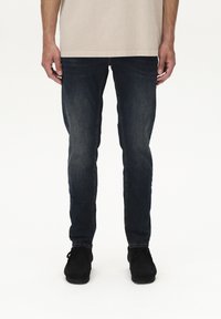 Tumši zili džinsa džinsi ar pieguļošu dizainu, kam piemīt izbalējusi tekstūra un smalkas šuvju detaļas, kombinēti ar melniem apaviem.