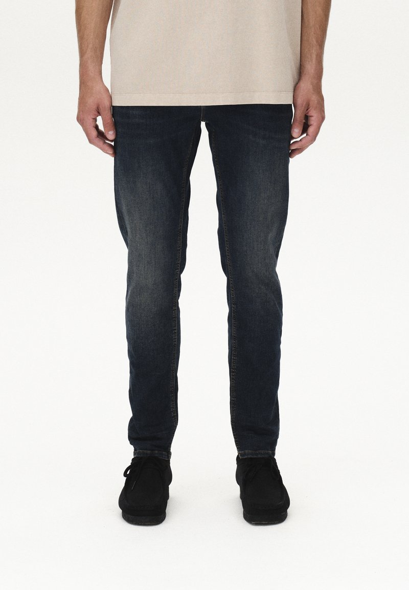 Tumši zili džinsa džinsi ar pieguļošu dizainu, kam piemīt izbalējusi tekstūra un smalkas šuvju detaļas, kombinēti ar melniem apaviem.