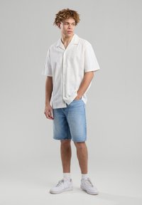 White short-sleeve shirt in a textured pattern, paired with light blue denim shorts. Model wears white sneakers with ankle socks.