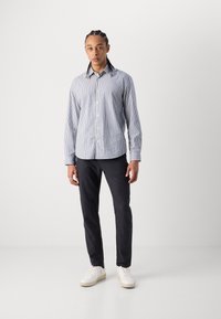 Blue and white striped button-up shirt with long sleeves, paired with black pants and white sneakers, standing against a plain background.