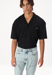 Chemise noire à manches courtes avec boutons, dotée de deux poches avant et d'un tissu texturé. Portée avec un jean en denim délavé au lavage léger, offrant une coupe décontractée.
