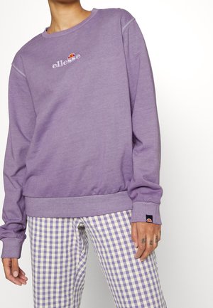 Persona que lleva un sudadera de ellesse color lavanda y pantalones de cuadros morados y blancos, mostrando dedos tatuados en la mano derecha.