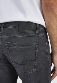 Ciemnoszare dżinsy o gładkiej fakturze, z czarną skórzaną naszywką z logo na tylnej części pasa i kwadratowym wzorem przeszycia kieszeni.