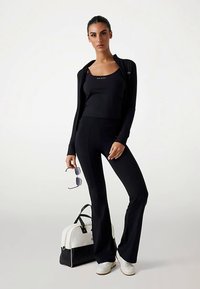 Musta urheiluasu, joka sisältää istuvan pitkähihaisen takin, toppitopin ja liehuvat housut. Asusteisiin kuuluvat aurinkolasit ja mustavalkoinen laukku.