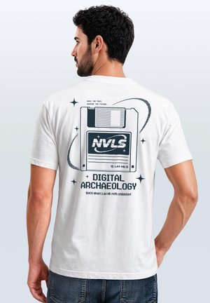 Man with dark hair wearing a white T-shirt featuring a large floppy disk graphic and text about digital archaeology on the back.