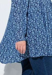 Niebieska bluzka z długim rękawem z wzorem jasnoniebieskich margaretek, wykonana z miękkiego materiału, o rozkloszowanym kroju i zaokrąglonym dole.