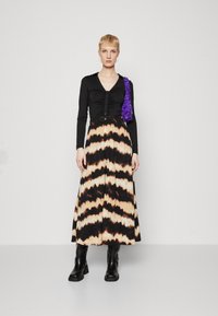 Dlhá čierna a béžová sukňa s tie-dye vzorom maxi s priliehavým čiernym topom s gombíkmi a dlhými rukávmi. Doplnkovaná fialovou taškou a čiernymi čižmami.