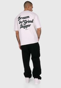 White cotton t-shirt featuring a large black text design on the back reading "Dream Big, Grind Bigger." Worn with black pants and white sneakers.
