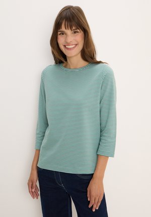Femme souriante avec des cheveux bruns portant un haut à rayures teal et blanches à manches trois-quarts et un jean bleu foncé, se tenant contre un mur blanc.