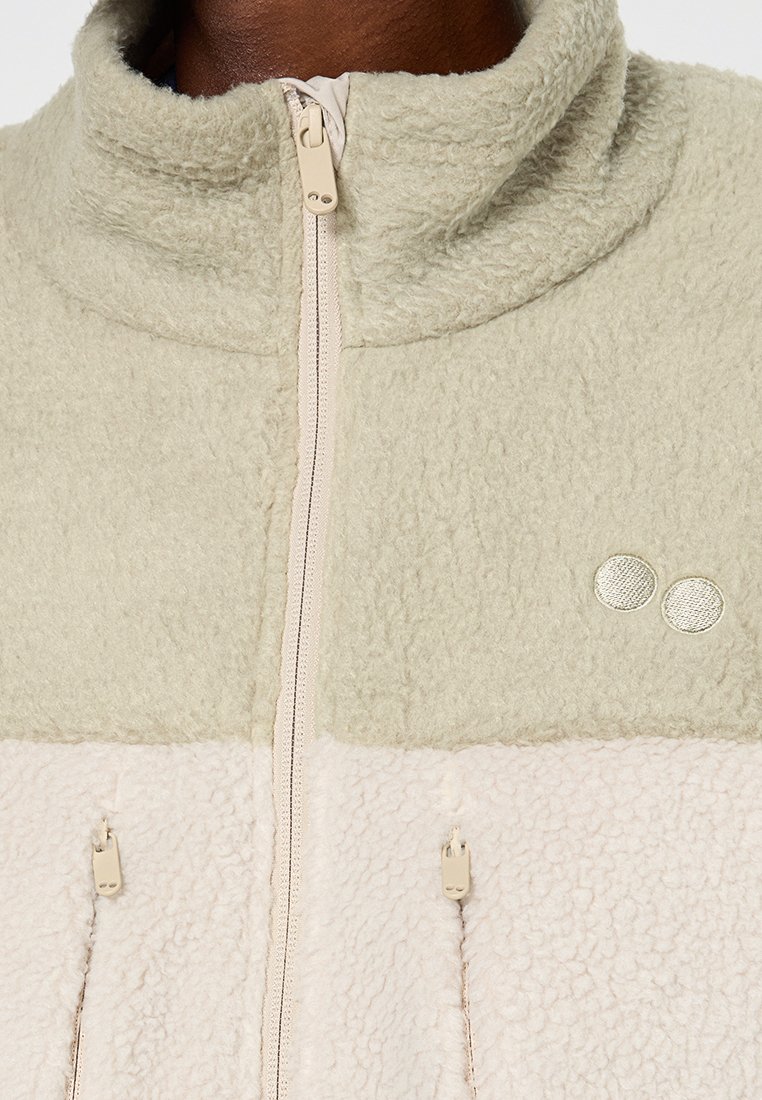 Beige und cremefarbene Fleecejacke mit durchgehendem Reißverschluss, hohem Kragen, zwei seitlichen Reißverschluss-Taschen und zwei kleinen runden Logopatches auf der Brust.